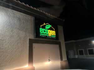 A building exterior at night with a lit sign reading "Ecogen Pest Control" above closed double doors. The sign features a green leaf graphic and house outline, highlighting Ecogen's commitment to environmentally friendly solutions.