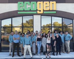 A group of adults and one child pose for a photo in front of a building with a large Ecogen sign. Most people wear matching blue Ecogenpest shirts and khaki pants, while a few are dressed casually.
