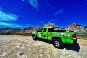 An Ecogen green pickup truck with company branding and a phone number is parked on a dirt road, framed by rocky mountains and a blue sky with clouds in the background.