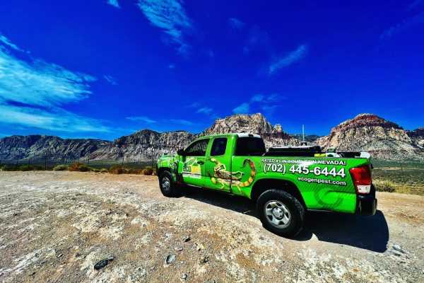 An Ecogen green pickup truck with company branding and a phone number is parked on a dirt road, framed by rocky mountains and a blue sky with clouds in the background.