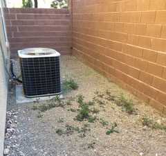 An Ecogen air conditioning unit sits on a concrete pad next to a house, surrounded by gravel and sparse weeds. A brick wall runs along the right side and back of the enclosed area.